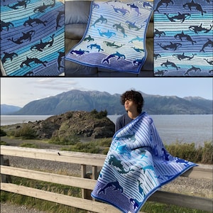 May include: A blue and white crocheted blanket with a pattern of orcas. The blanket is draped over a wooden railing and a person is sitting on a rock in the background.