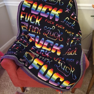 May include: A black crocheted blanket with the word "FUCK" in rainbow colors. The blanket is draped over a red armchair. The blanket has a purple border.