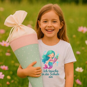 May include: A young girl smiles, holding a large, cone-shaped school bag. The bag is pastel pink, mint green with white polka dots, and beige. She wears a white t-shirt with a mermaid graphic and the text "Ich tauche in die Schule ein."