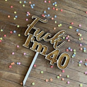 Könnte beinhalten: Holz-Kuchentopper mit dem Text "F*ck I'm 40" in brauner Holzfarbe. Der Topper befindet sich auf einer Holzoberfläche mit bunten Streuseln.