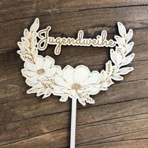 May include: A wooden cake topper featuring a floral wreath and the text "Jugendweihe" in a cursive font.