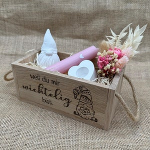 Könnte beinhalten: Eine Holzkiste mit Henkel, gefüllt mit einer rosa Kerze, einem weißen herzförmigen Kerzenhalter, einer weißen Gnomfigur und Trockenblumen. Die Kiste hat den Text "weil du mir wichtig bist" auf der Vorderseite eingraviert.