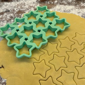 May include: Green star-shaped cookie cutters on top of a sheet of cookie dough with star-shaped cutouts.
