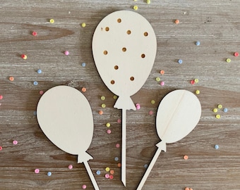 Może przedstawiać: Trzy drewniane kształty balonów z przymocowanymi patyczkami. Balony są gładkie, z wyjątkiem jednego, który ma małe otwory na całej powierzchni. Balony znajdują się na drewnianym tle z kolorowymi konfetti.