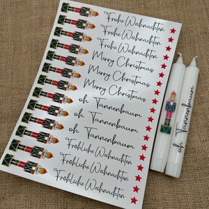 Può includere: Un foglio di carta argentata con più immagini di schiaccianoci e saluti di Natale in tedesco e inglese. I saluti includono "Frohe Weihnachten" e "Merry Christmas". Ci sono anche candele bianche con un'immagine di schiaccianoci e il testo "oh Tannenbaum".