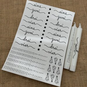 Puede incluir: Un conjunto de pegatinas blancas para calendario de adviento con números negros y palabras alemanas para los números del uno al cuatro. Las pegatinas están sobre un fondo plateado. También hay pequeñas pegatinas de árboles de Navidad negros. Hay tres velas blancas con números negros en ellas.