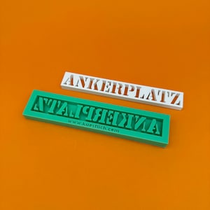 Puede incluir: Dos moldes de plástico blanco para hacer chocolate u otras golosinas. El molde superior está etiquetado como "ANKERPLATZ" y el molde inferior está etiquetado como "ANKERPLATZ" al revés. El molde inferior también tiene el sitio web "www.kuestlich.com" impreso en él.