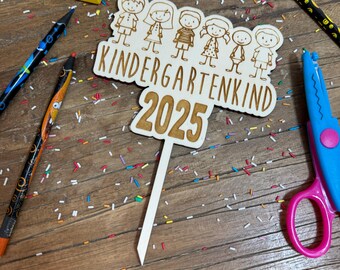 Könnte beinhalten: Ein Holzschild mit dem Text "Kindergartenkind 2025" und einer Reihe von Strichmännchen. Das Schild ist auf einem Stock und von bunten Streuseln umgeben.