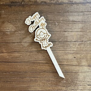 May include: A white wooden cake topper with the words "Happy Birthday" and a cartoon girl with a ponytail and a dotted dress.