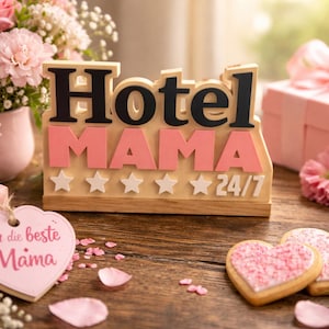 以下が含まれることがあります： 黒とピンクの文字で「Hotel MAMA 24/7」と書かれた木製の看板。星のアクセント付き。ピンクの花、ハート型のクッキー、ピンクのリボンが付いたギフトボックスが周りに配置されています。