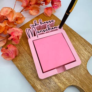 Könnte beinhalten: Ein rosa Notizblockhalter mit dem deutschen Text "Ich hasse Menschen" (Ich hasse Menschen). Der Halter hat ein quadratisches Design mit einem Schlitz für einen Stift. Ein rosa Haftnotizblock befindet sich im Inneren. Der Halter befindet sich auf einem Holzschneidebrett.