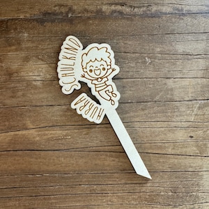 May include: A white wooden stick with a cutout of a smiling child jumping with the words "Hurra" and "Schulkind" around the child.