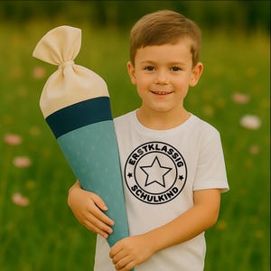 Peut inclure: Un enfant tient un grand sac d'école en forme de cône. Le sac est crème, bleu sarcelle et bleu marine. L'enfant porte un t-shirt blanc avec un graphisme circulaire noir qui dit "ERSTKLASSIG SCHULKIND" et une étoile.