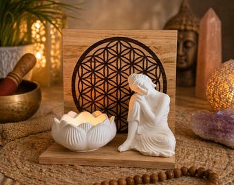 May include: A white ceramic lotus flower candle holder with a lit candle, a white Buddha statue, and a wooden block with a geometric design. A wooden prayer bead necklace is in the foreground. The theme is spiritual and meditative.