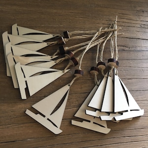May include: A group of wooden sailboat decorations. Each ornament has a light-coloured sail with a cut-out design and a dark brown wooden base. They are strung together with natural twine, ideal for nautical-themed decor.