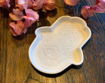 May include: White ceramic dish shaped like a hamsa hand with intricate details. The hamsa is a symbol of protection and good luck.