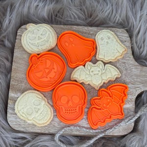Könnte beinhalten: Ein Holzbrett zeigt Halloween-Kekse und orangefarbene Ausstecher. Die Kekse sind in Form eines Kürbisses, Geistes, Schädels und einer Fledermaus. Die orangenen Ausstecher haben die gleichen Formen. Der Hintergrund ist ein weiches, graues, pelziges Material.