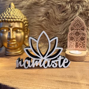 Könnte beinhalten: Eine goldene Buddha-Statue, ein schwarz-weißes Lotusblumen-Design und ein hölzerner Kerzenhalter mit floralem Muster werden auf einer Holzoberfläche ausgestellt. Das Wort "namaste" ist in weißer Schrift unter der Lotusblume geschrieben.