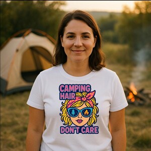 May include: A woman wearing a white t-shirt with the slogan "CAMPING HAIR DON'T CARE". The graphic shows a cartoon woman with sunglasses, a pink bandana, and blonde hair. A tent and campfire are visible in the background.