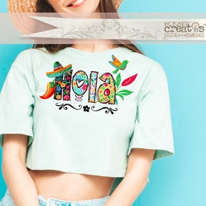 May include: A light mint green short-sleeved crop top featuring a colorful graphic print of the word Hola. The letters are filled with floral patterns and accompanied by a small sombrero, a chili pepper, a bird, and decorative black swirls.