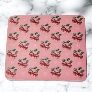 May include: Pink rectangular mousepad featuring a repeating pattern of red cherries with green stems and leaves. The cherries have a glossy appearance. Small white bows are scattered across the background. The text "The Mamas" is printed on the mousepad.