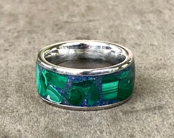 May include: A silver-toned metal ring featuring a wide band inlaid with a mosaic of green malachite stone fragments and deep blue lapis lazuli accents. The polished metal edges frame the colorful stone inlay, creating a smooth, continuous circular band with a reflective interior surface.