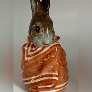 May include: A ceramic figurine of a brown rabbit wrapped in an orange blanket with white stripes and small white dots. The rabbit has long ears, dark eyes, and a smooth, glossy finish. The figure is depicted in a seated position, fully covered.