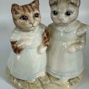 May include: A ceramic figurine featuring two anthropomorphic cats standing side-by-side. The cat on the left has brown tabby markings, while the cat on the right is grey. Both cats wear pale blue dresses with collars and stand on a textured base.