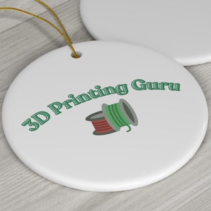 May include: Two white, round ceramic ornaments with gold string hangers. One ornament features the text "3D Printing Guru" and an illustration of two spools of filament. The other ornament is blank.