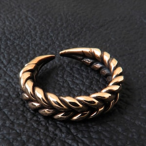 May include: A gold-toned ring with a braided design. The ring is open and has a pointed end.
