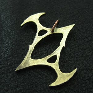 May include: A brass-coloured pendant with a unique, stylised design. The pendant features a central open oval shape, with pointed edges and curved accents. A small copper-coloured ring is attached at the top for hanging.