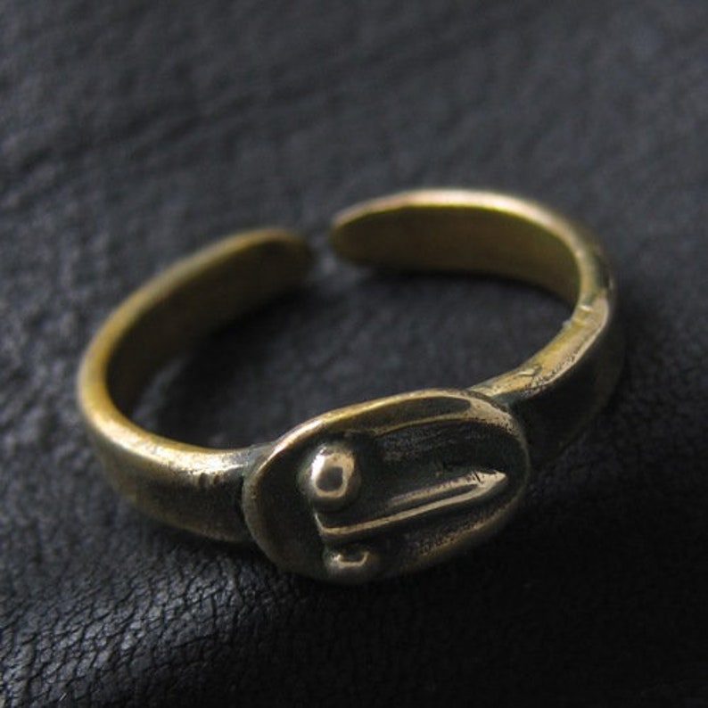 May include: A brass-coloured adjustable ring with an oval centrepiece. The oval has a stylized design with a central line and a small dot. The ring has a vintage or antique appearance.