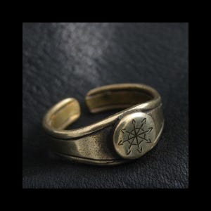 May include: A bronze-colored adjustable ring with an engraved starburst design on a circular plate. The ring has a slightly weathered appearance, with a band that wraps around the finger and an open design.