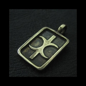 Puede incluir: Colgante rectangular con un diseño estilizado. El colgante tiene un fondo oscuro y texturizado con un borde más claro y en relieve. El diseño central presenta una forma de cruz con elementos curvos y un lazo para una cadena.