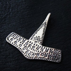 May include: A silver-toned pendant with a unique, geometric design. The pendant features a central cross-like pattern and is adorned with small, circular indentations. The piece has a textured appearance and is set against a dark background.