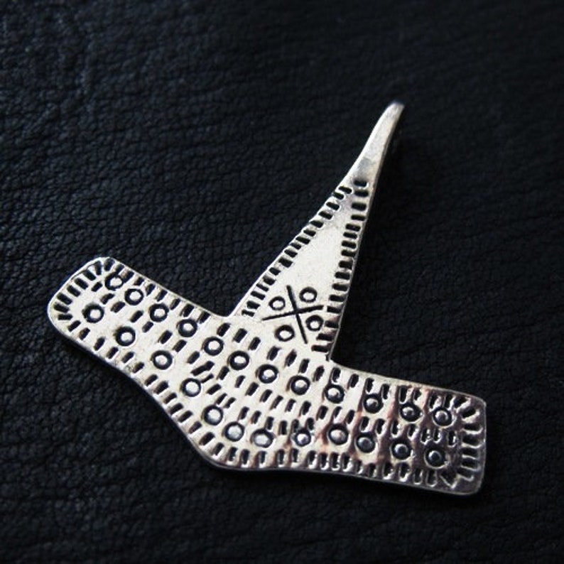 May include: A silver-toned pendant with a unique, geometric design. The pendant features a central cross-like pattern and is adorned with small, circular indentations. The piece has a textured appearance and is set against a dark background.