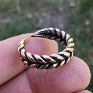 Puede incluir: Un anillo de color bronce con un dise&ntilde;o de cuerda trenzada. El anillo est&aacute; abierto en la parte superior y tiene un extremo puntiagudo. El anillo se sostiene en una mano sobre un fondo verde borroso.