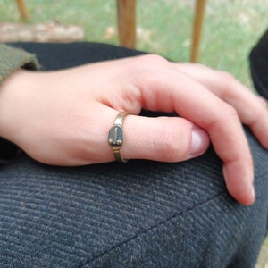 May include: A gold-coloured ring with a rectangular design on the thumb. The ring has a dark, oval-shaped design in the centre. The person is wearing a green tweed jacket. The ring is worn on a person's thumb.