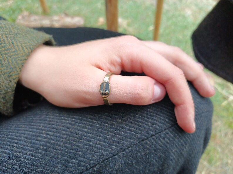 May include: A gold-coloured ring with a rectangular design on the thumb. The ring has a dark, oval-shaped design in the centre. The person is wearing a green tweed jacket. The ring is worn on a person's thumb.