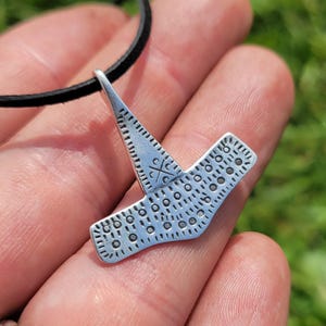 May include: A silver-colored pendant in the shape of a hammer, with a black leather cord. The hammer has a detailed design with lines and dots. The pendant is held in a person's hand.