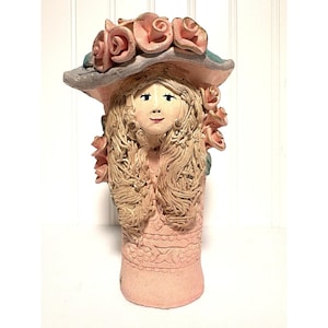 Puede incluir: Un jarrón de cerámica con forma de persona que lleva un sombrero de ala ancha adornado con rosas rosadas. La figura tiene cabello texturizado de color claro, un rostro pintado con ojos negros y una boca pequeña, y una base rosa con patrones florales en relieve.