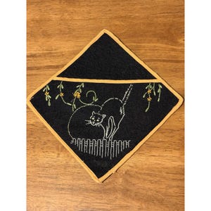 May include: A diamond-shaped black fabric potholder with a yellow border. The front features white embroidered line art of a cat standing on a fence, surrounded by green vines with small yellow flowers. The design is centred on the fabric.