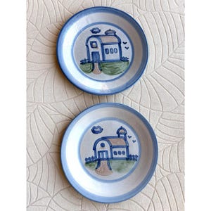 May include: Two ceramic plates with a hand-painted design. Each plate features a blue rim and a central image of a house with a blue roof, green grass, and a blue fence. The plates are on a cream-colored background.