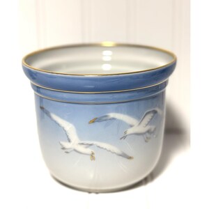 May include: A ceramic planter with a light blue band and gold trim. The planter features a design of white seagulls in flight against a light blue sky. The interior is white.