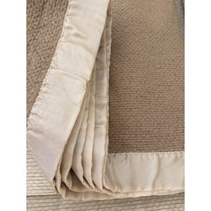 K&ouml;nnte beinhalten: Eine gefaltete beige Decke mit einem cremefarbenen Satinrand. Die Decke hat ein strukturiertes Gewebe, und der Satinrand ist glatt und gl&auml;nzend. Die Decke ist gefaltet und zeigt den Kontrast zwischen den Texturen und Farben.