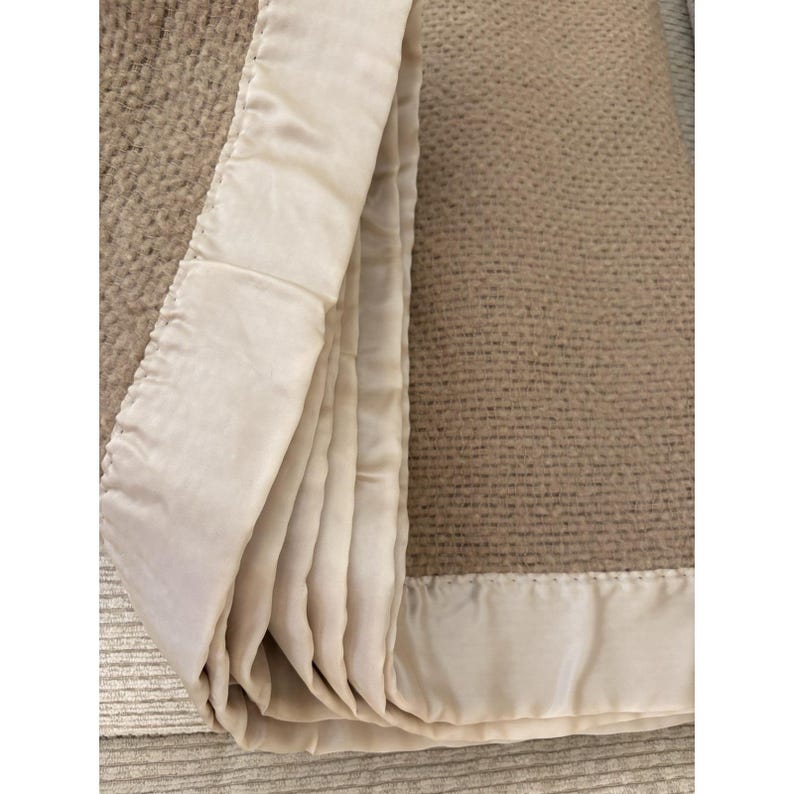 K&ouml;nnte beinhalten: Eine gefaltete beige Decke mit einem cremefarbenen Satinrand. Die Decke hat ein strukturiertes Gewebe, und der Satinrand ist glatt und gl&auml;nzend. Die Decke ist gefaltet und zeigt den Kontrast zwischen den Texturen und Farben.