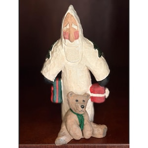 May include: A carved wooden Santa Claus figurine in a white robe with green and black accents, holding a red and green gift bag and a red mitten. A small brown teddy bear with a green scarf sits in front of Santa.
