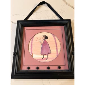 May include: A framed artwork featuring a girl in a pink dress holding flowers, set against a pink background within a circular frame. The artwork is enclosed in a black frame with a black ribbon for hanging.