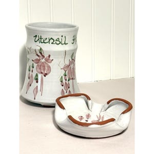 Puede incluir: Un soporte para utensilios de cerámica blanca con las palabras "Utensil Holder" en escritura verde. El soporte presenta una forma acampanada y está decorado con diseños florales rosas y verdes. También hay un reposacucharas de cerámica blanca a juego con un borde marrón.