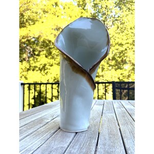 Peut inclure: Un vase en céramique bleu clair avec un design incurvé unique. Le bord et les bords présentent un accent marron, ajoutant du contraste à la surface lisse et brillante. Le vase mesure environ 25 cm de haut.